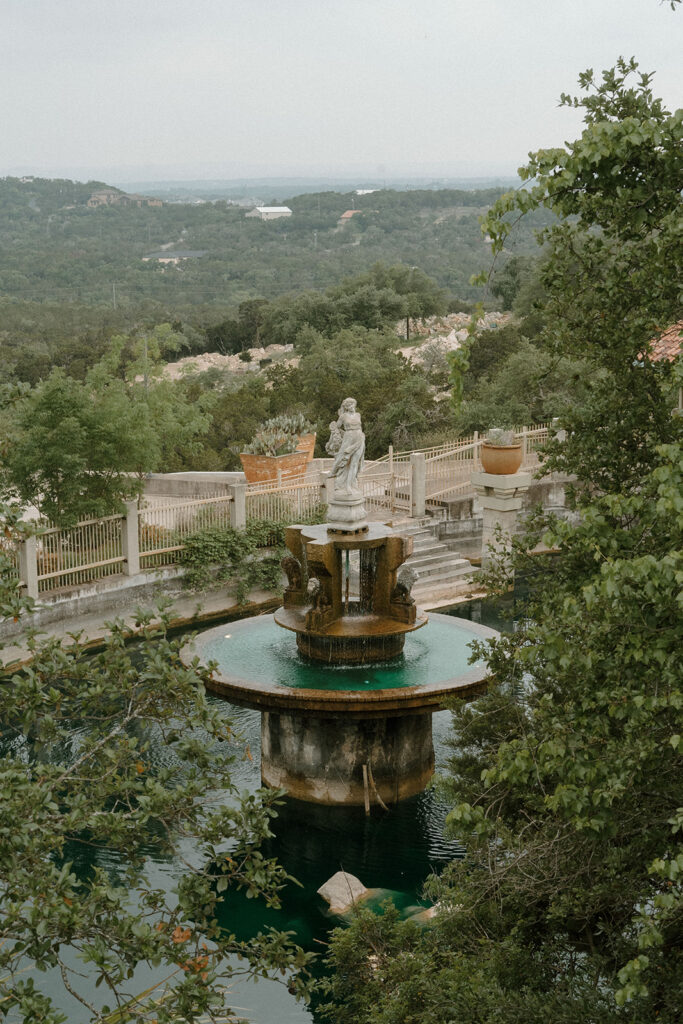 Elevated view of a tiered stone fountain topped with a classical statue, surrounded by lush greenery and overlooking rolling Texas Hill Country hills in the distance.