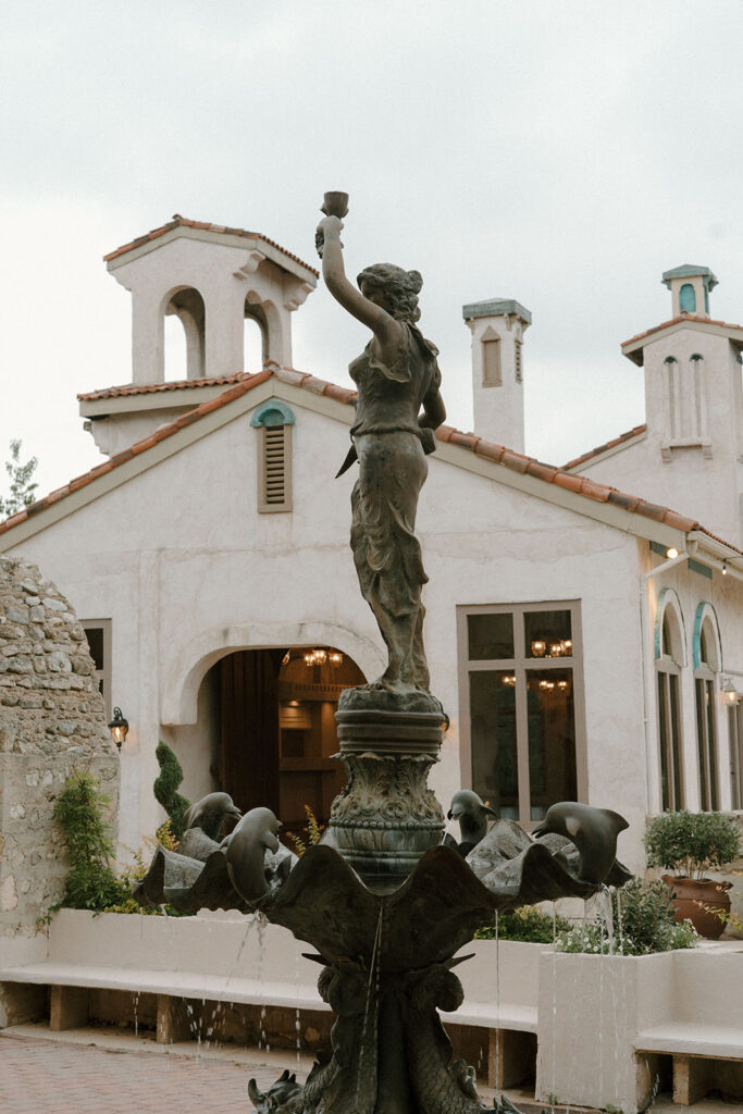 Bronze fountain statue of a woman holding a cup aloft, surrounded by sculpted dolphins, set in front of a Mediterranean-style villa with arched windows and terracotta roof tiles