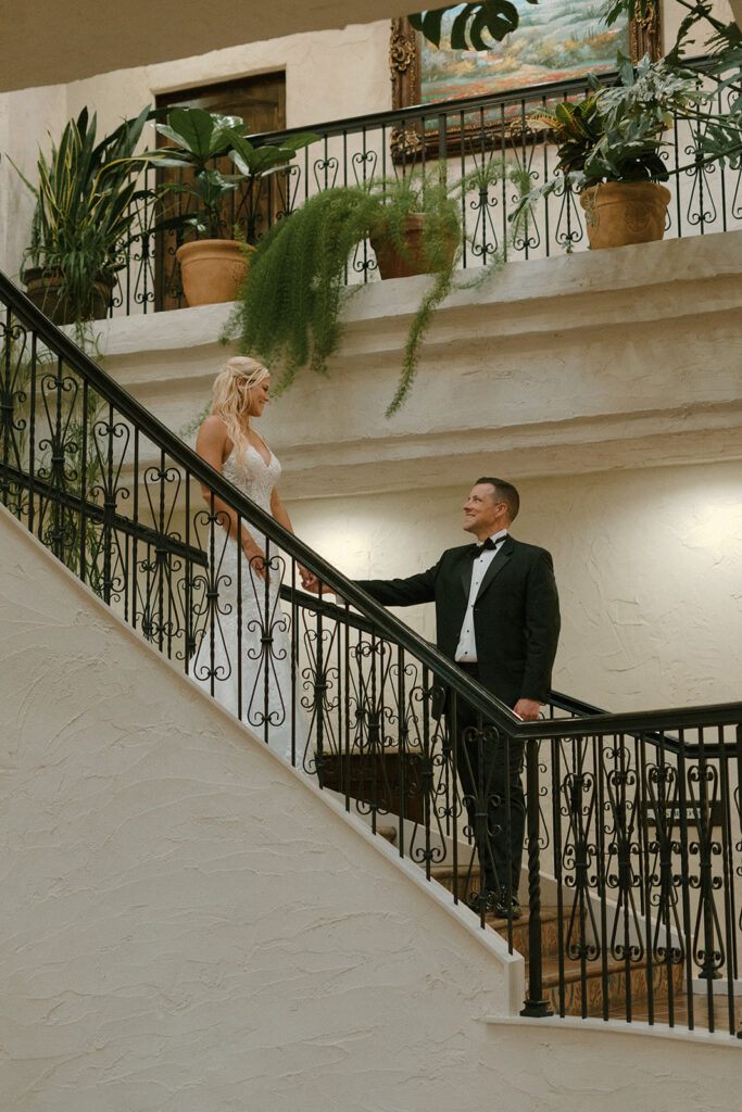Bride in a lace gown descending a grand staircase lined with wrought-iron railings, holding hands with the groom in a tuxedo, surrounded by lush potted greenery.  