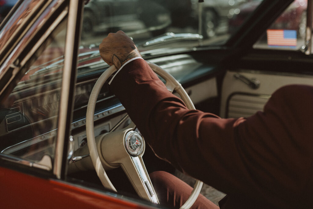 a groom's arm in a maroon suit in a red vintage car