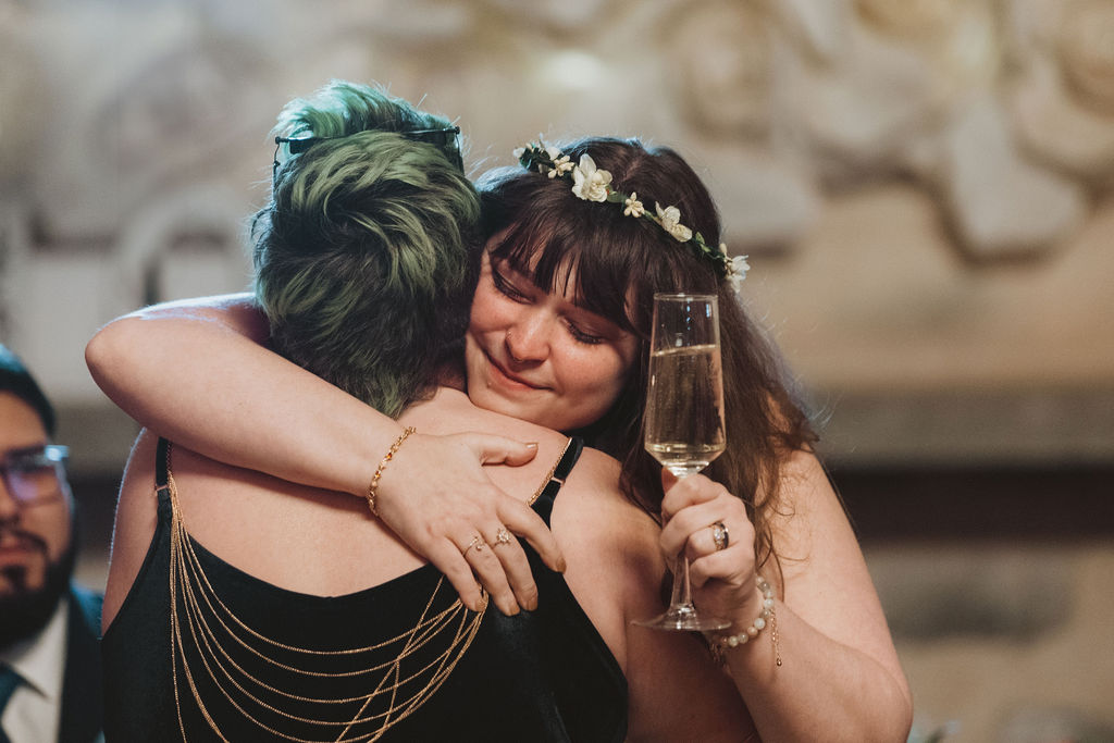 Two friends embrace tightly in an emotional moment while the bride holds a glass of champagne