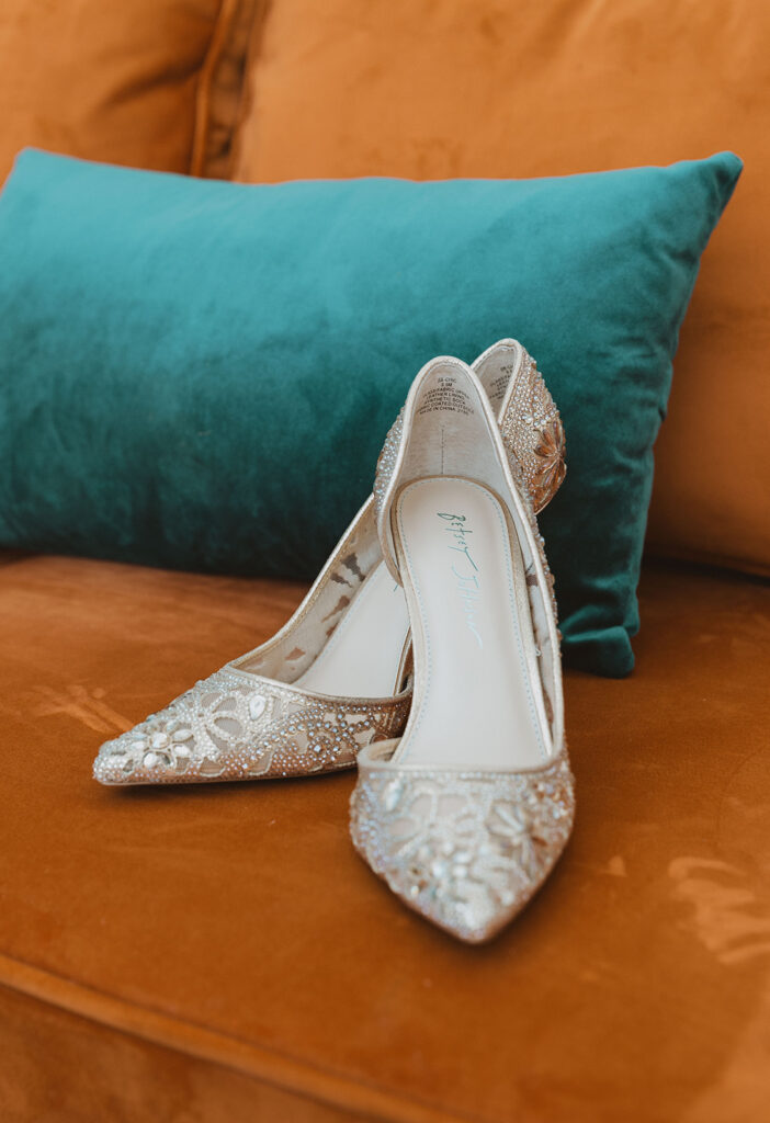 golden bedazzled shoes branded by Betsy Johnson on a bright orange couch with a teal throw pillow behind it