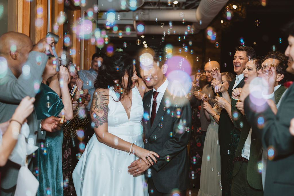 A bride and groom walk through bubbles surrounded by loved ones