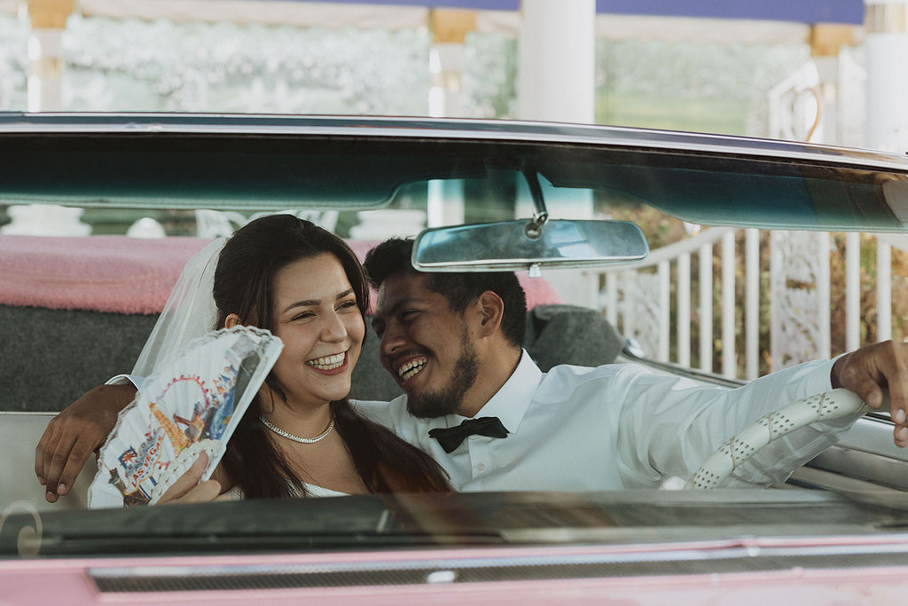 A bride and groom sit close together behind the wheel of a pink cadilac