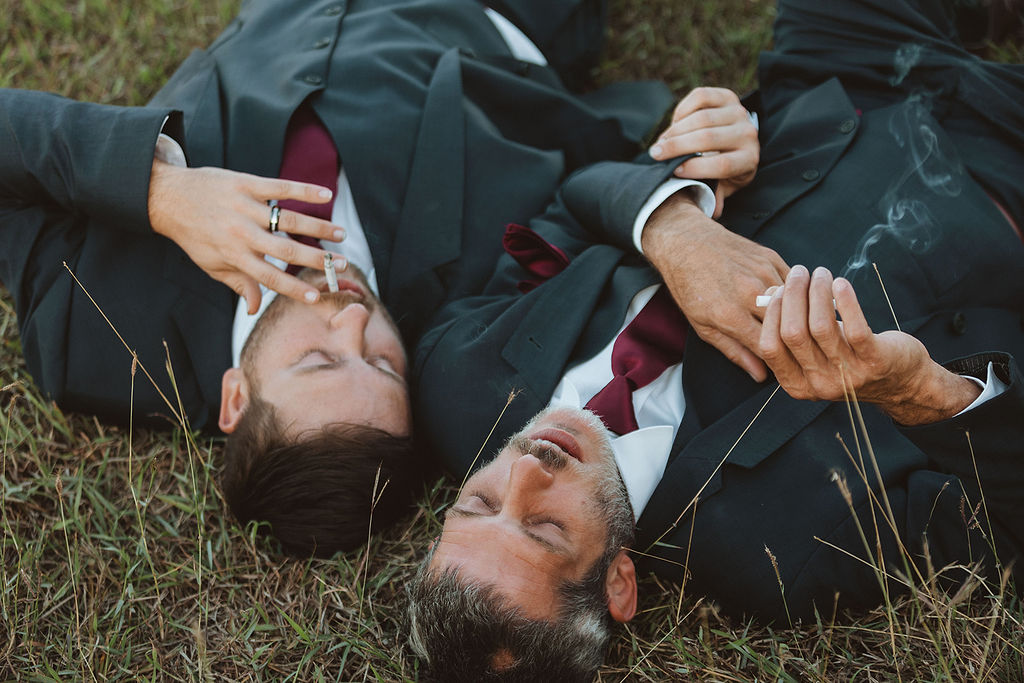 Two grooms lay down in a field and smoke a cigarette